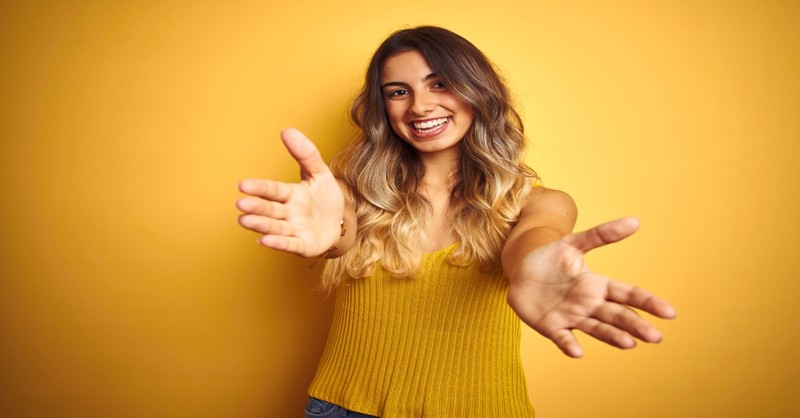 Young woman w/ yellow background looking at camera smiling with open arms for hug, love my neighbor